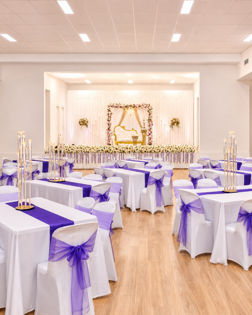 Function hall styled with white linen, purple sashes and a floral stage